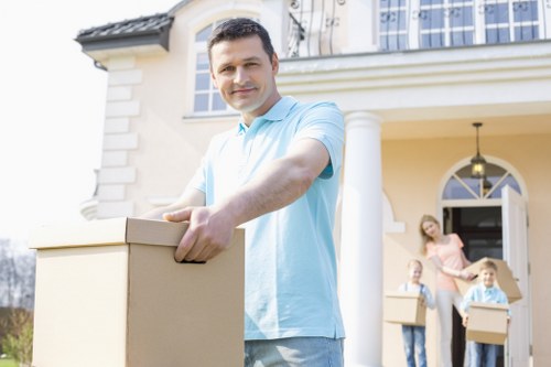 A man wearing a light blue shirt stands outside a residential house in Shepherds Bush, holding a medium-sized cardboard box in his hands and smiling towards the camera. Behind him, a family of three, including a woman and two children, are seen carrying additional cardboard boxes, indicating a house move. The house features a cream-colored exterior with decorative white moldings, a prominent arched window, and a small porch with a black lantern-style light fixture. The scene suggests an outdoor transfer of household belongings, with the family preparing to load or unload items either at the front entrance or driveway, consistent with house removal services in the Shepherds Bush area. Natural daylight illuminates the scene, highlighting the cardboard boxes' brown matte finish, the varied textures of the family’s clothing, and the house’s clean, well-maintained facade, subtly reinforcing local residential removal services provided by Shepherds Bush Man with Van.